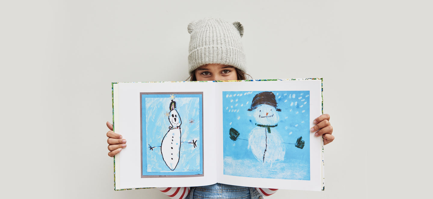 A child in a winter wooly hat holds a kids' art keepsake book open showing two children's drawings of snowmen