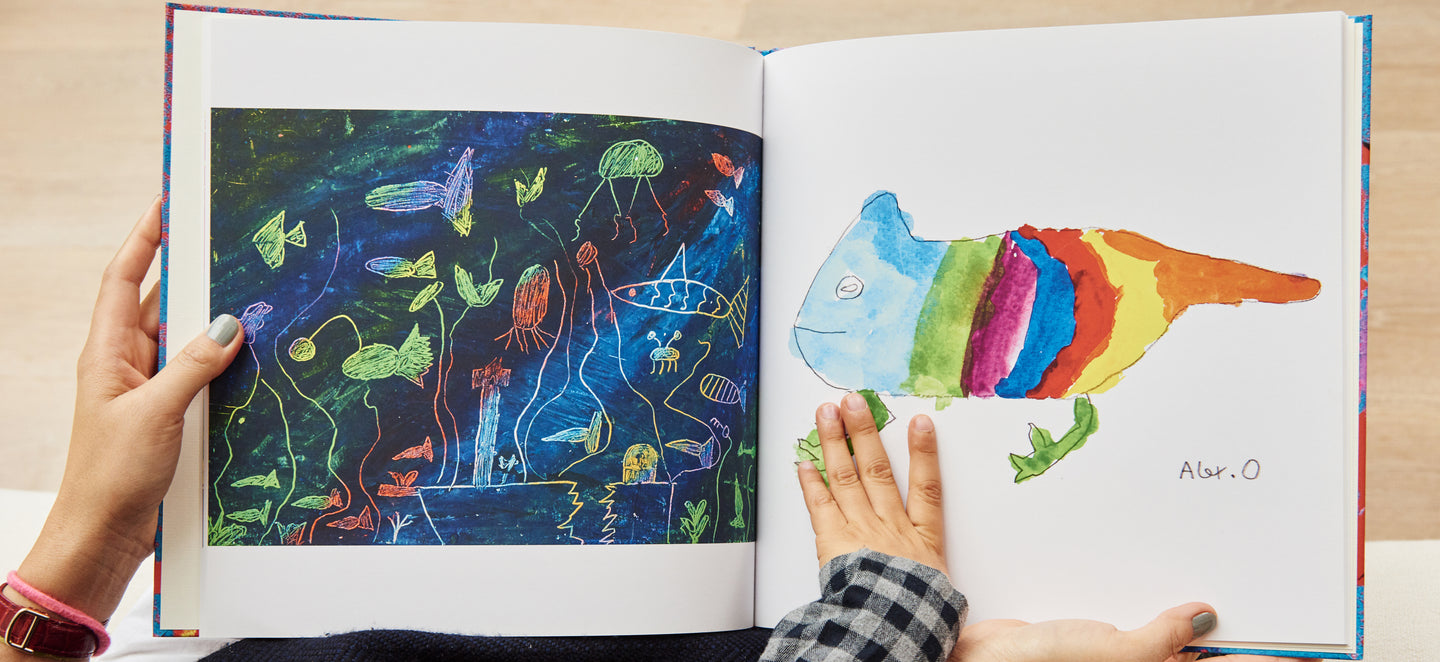 Mum holds a book open with kids' drawings in it while a child's hand touches the page
