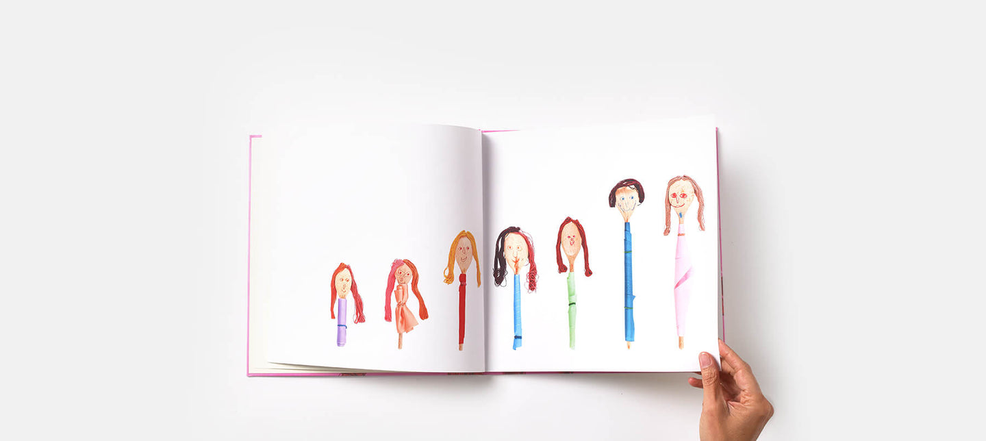 A hand holds a keepsake kids' art book open showing spoon puppets all standing in a line like a family