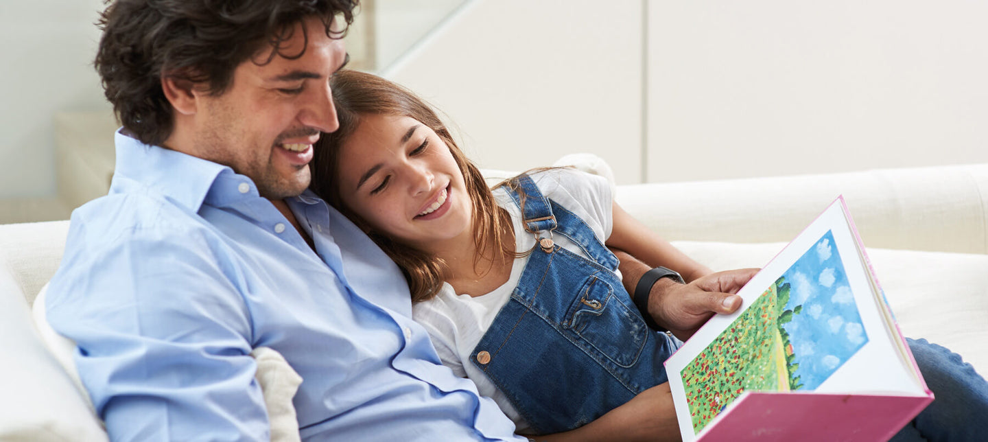 A dad and his daughter cuddle on a sofa and look through a book of her artwork