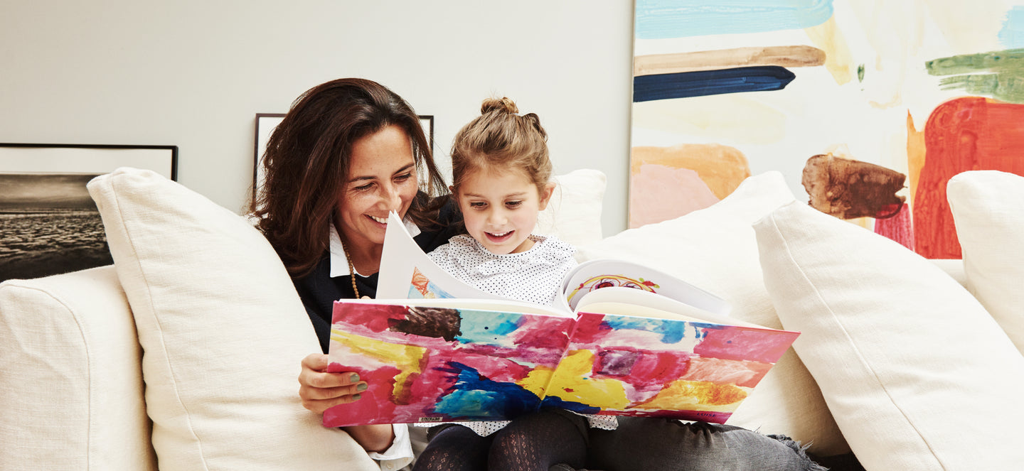 A mother and child sit on a sofa looking through a keepsake book of the daughter's artwork