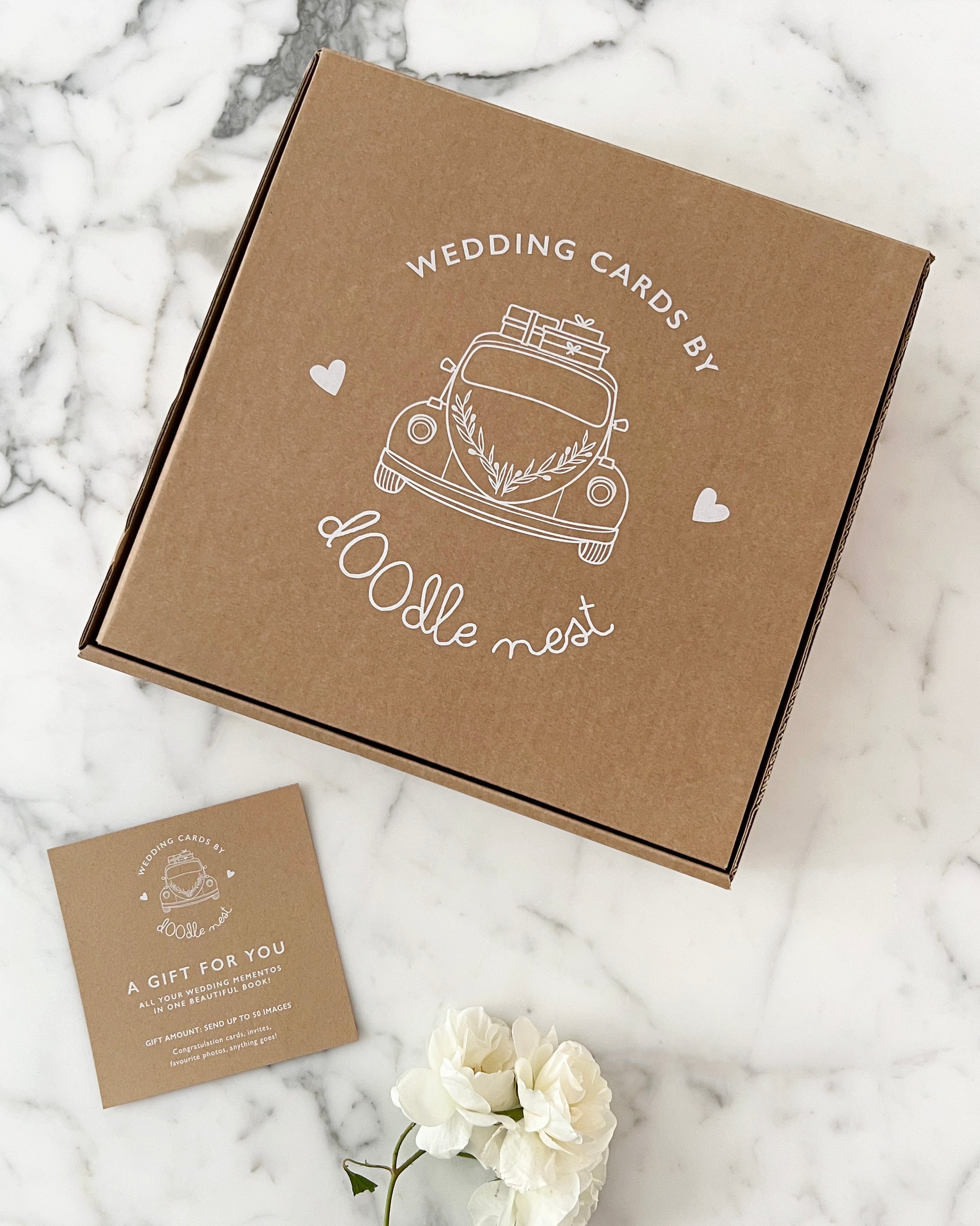 A kraft cardboard box with a graphic of a vintage car and hearts and the text Wedding Cards by Doodle Nest, alongside a gift voucher on a marble surface.