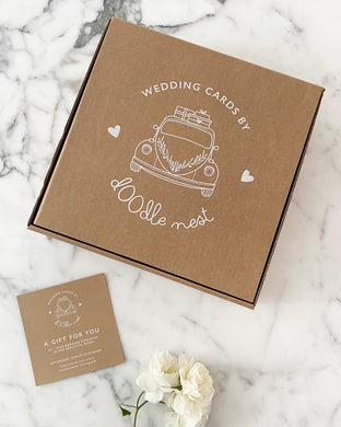 A kraft cardboard box with a graphic of a vintage car and hearts and the text Wedding Cards by Doodle Nest, alongside a gift voucher on a marble surface.