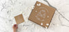 A kraft cardboard box with a graphic of a vintage car and hearts and the text Wedding Cards by Doodle Nest, alongside a gift voucher on a marble surface with white flower petals scattered around like confetti.