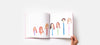 A hand holds a keepsake kids' art book open showing spoon puppets all standing in a line like a family